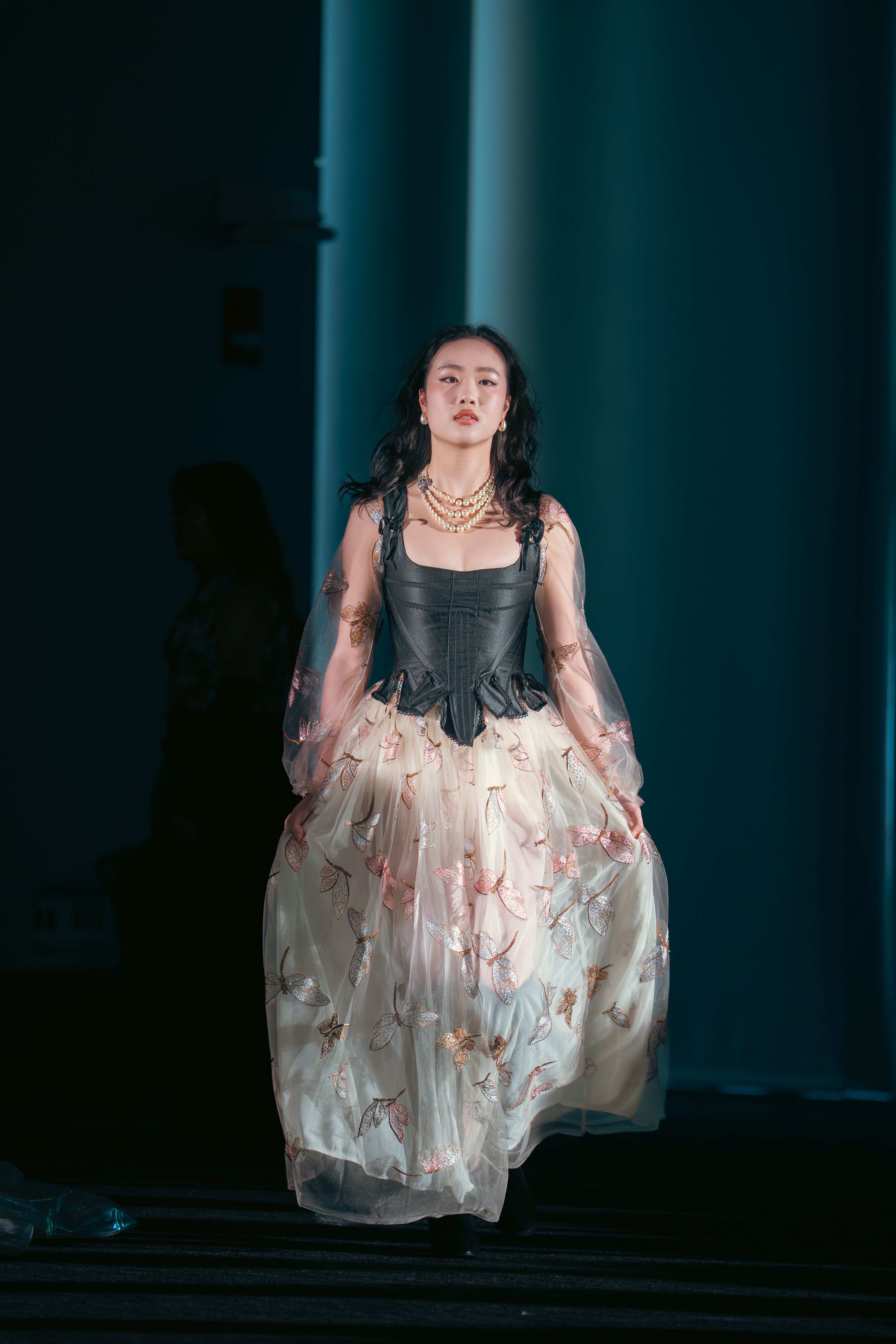 Model walks down the runway wearing a black corset-style top paired with a voluminous ivory tulle skirt embroidered with shimmering butterfly motifs in pink and gold. She accessorizes with layered pearl necklaces and wears her hair in soft waves.